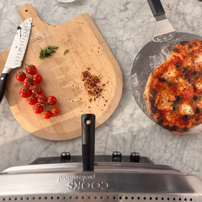 A top-down view of a stainless steel Cuisinart professional grill with a black handle, set on a gray marble countertop. Surrounding the grill are cooking ingredients and tools, including a wooden cutting board with cherry tomatoes, red pepper flakes, a chef's knife, and a pizza pan with a baked pizza.